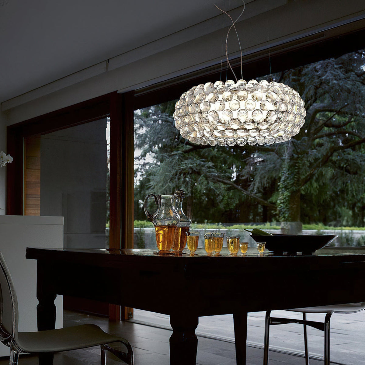 A clear sphere pendant hangs inside a dark wood room with glass doors to a garden, above a dark table with a jug of juice, from Clérachic.