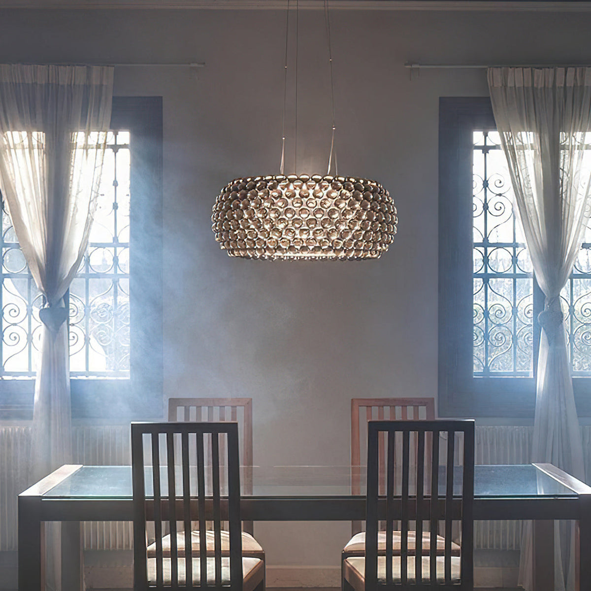 A sphere-covered pendant glows softly in a hazy room with sheer curtains and ironwork windows, above a glass table, from Clérachic.