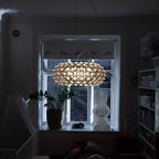 A sphere-covered pendant glows warm in a dim room, in front of a window with a plant and a bunny lamp, from Clérachic.