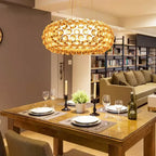A gold-amber sphere pendant glows above a wooden table laid for dinner, with a sofa and bookshelves behind, from Clérachic.