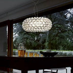 A clear sphere pendant glows in a room with a large window onto a tree, above a dark table with a jug and glasses, from Clérachic.
