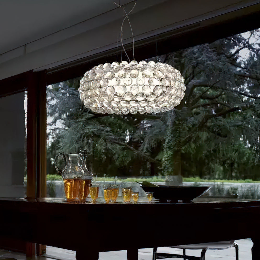 A clear sphere pendant glows in a room with a large window onto a tree, above a dark table with a jug and glasses, from Clérachic.