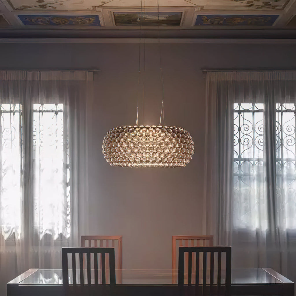 A sphere pendant glows warm between two tall curtained windows above a dark glass-topped table, from Clérachic.