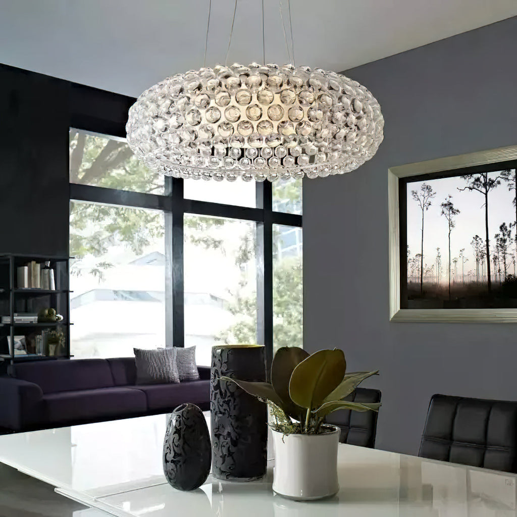 A clear sphere pendant hangs above a white table with a leafy plant and dark vases, in a grey room with large windows, from Clérachic.