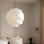 Clérachic globe pendant light lit above a bed with white pillows, cream bedding and a beige upholstered headboard, a bright patch showing where two bands part.