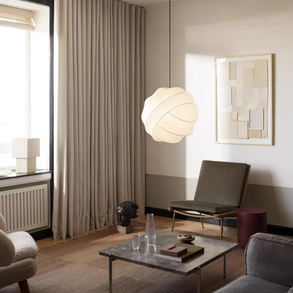 Clérachic globe pendant light lit between a curtained window and a framed paper collage, above a stone coffee table in a taupe-toned living room.