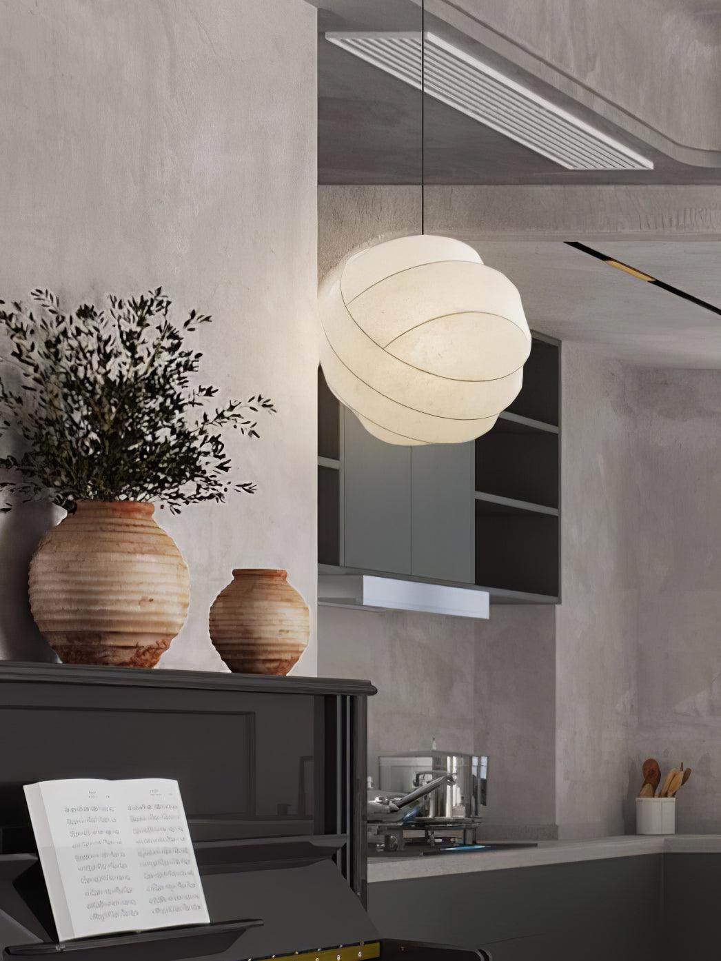 Clérachic globe pendant light lit towards a grey kitchen, seen past terracotta urns of olive branches standing on a dark piano.