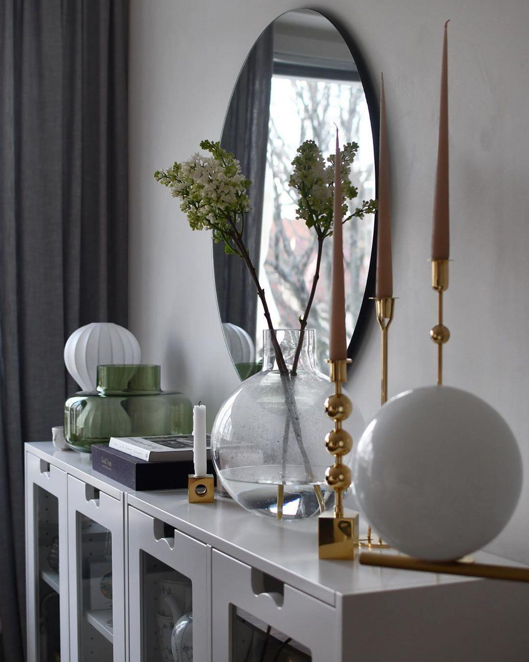 Clérachic lamp on a white glazed cabinet, with a round mirror, a glass vase of blossom and a brass candle holder.