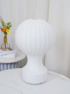 Front view of the larger Clérachic lamp, unlit, on a white marble table, with sheer curtains and a small vase of flowers behind.