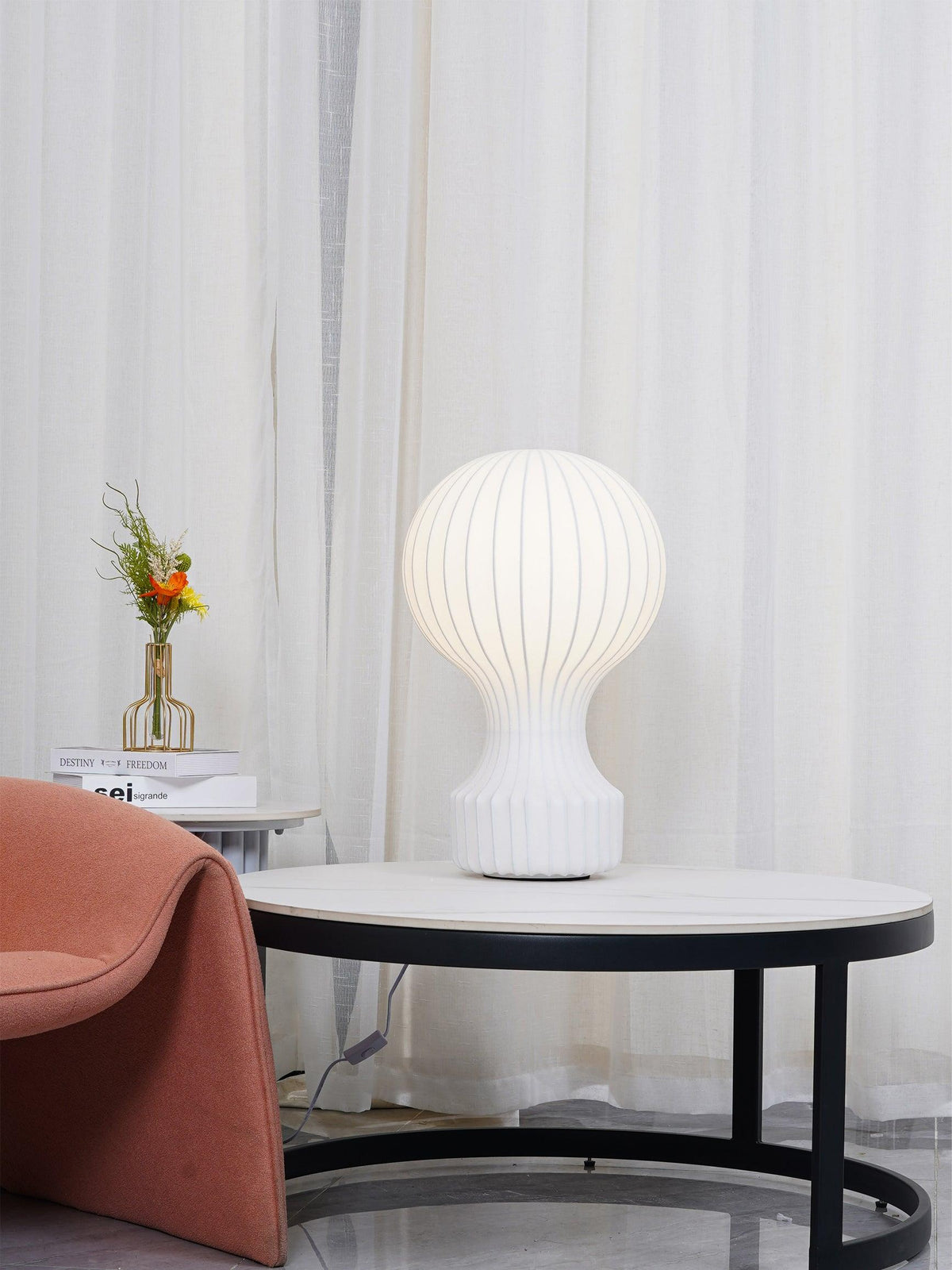 A Clérachic table lamp glowing on a round marble coffee table, with a coral armchair and a black metal frame in view.