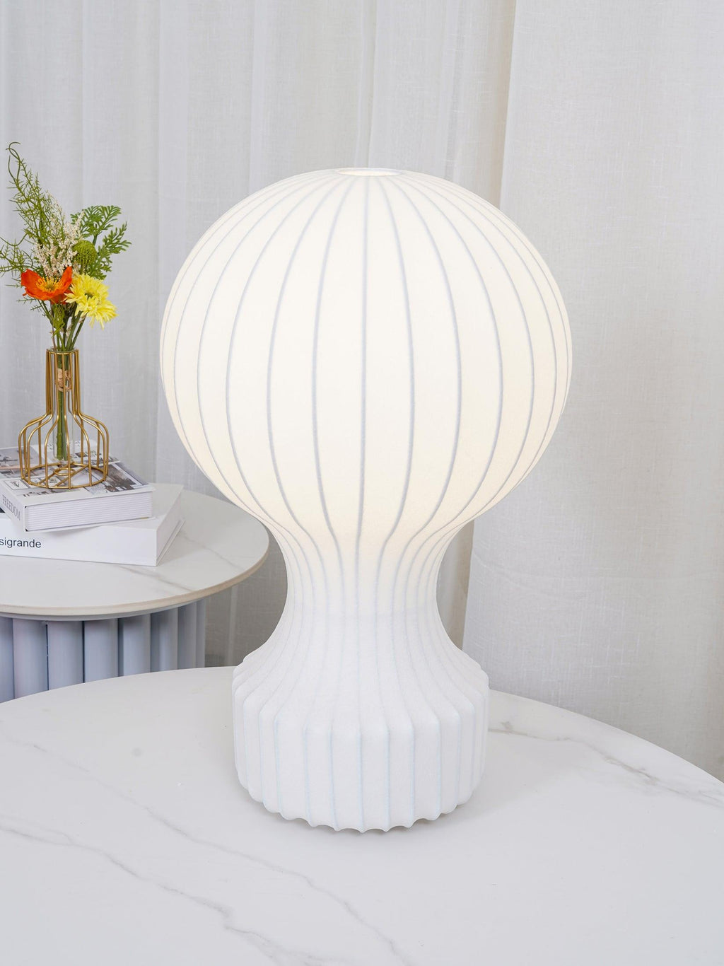 Close view of the lit Clérachic lamp on a marble table, its shade glowing evenly with fine ribs, and curtains behind.