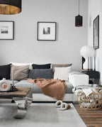 Clérachic lamp in the corner of a grey living room, behind a sofa with a tan throw and sheepskin slippers on the rug.