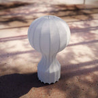 A Clérachic lamp outdoors on sunlit ground with dappled leaf shadows, its ribbed white base standing on the dirt.