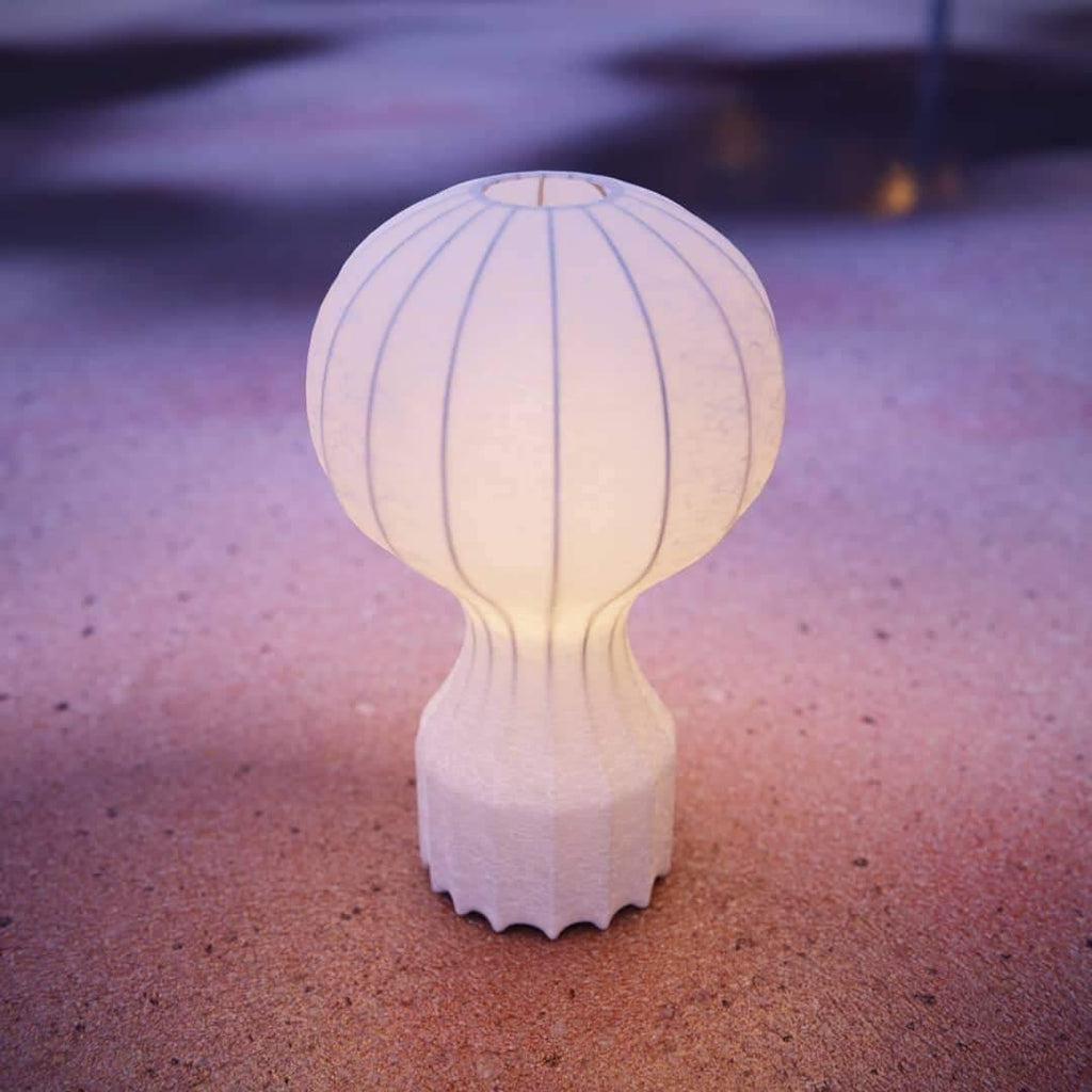 The Clérachic lamp lit with a warm glow, standing on pink and purple sand at dusk.