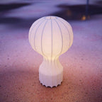 The Clérachic lamp lit with a warm glow, standing on pink and purple sand at dusk.