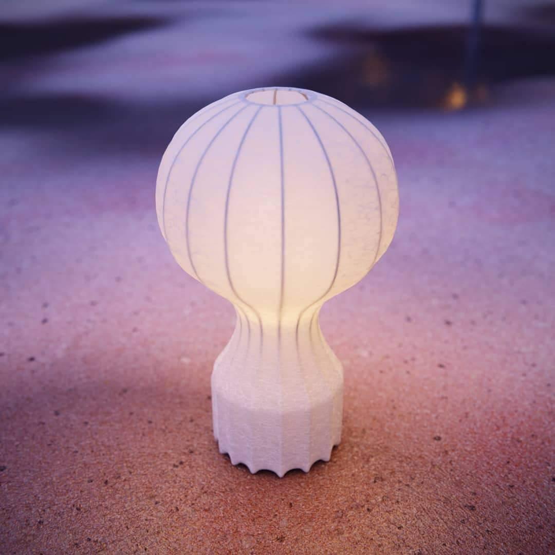 The Clérachic lamp lit with a warm glow, standing on pink and purple sand at dusk.