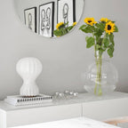 A Clérachic table lamp on a white sideboard beneath a round mirror, with sunflowers in a glass vase and a small candle tray.