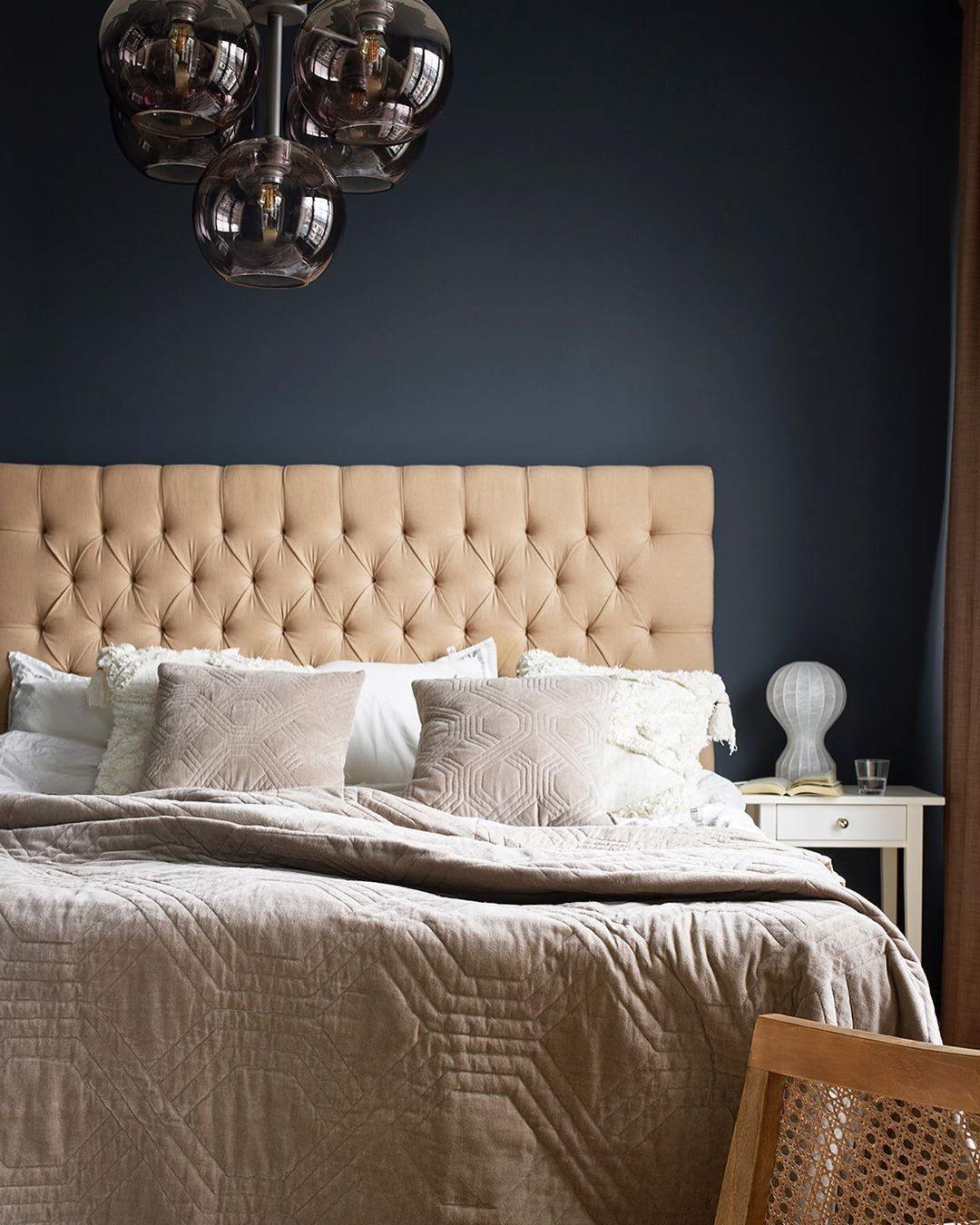 A bedside Clérachic lamp on a white nightstand in front of a navy wall, with a tufted headboard and a smoked-glass pendant.