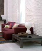 A Clérachic lamp on a corner of a low dark table, beside a nest-shaped bowl and a plum sofa in front of white brick.