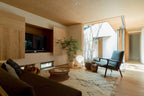 A distant, lit Clérachic lamp on the floor of a timber-panelled living room, with a green armchair and a woven rug.