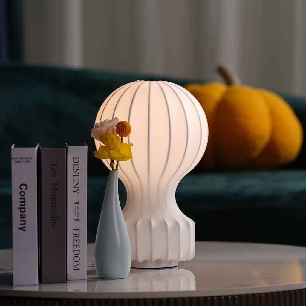 Close view of a Clérachic lamp glowing warmly beside a tall pale-blue vase, three books and an orange pumpkin cushion.