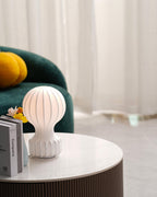 Clérachic table lamp glowing on a marble coffee table, with a green sofa and an orange cushion at the left edge.