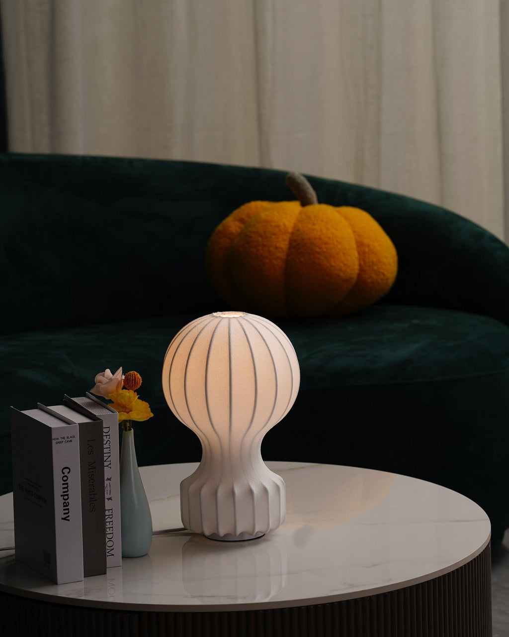 A lit Clérachic lamp on a round marble table in front of a green sofa, with books and a small vase at its side.