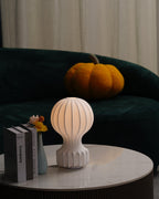 A lit Clérachic lamp on a round marble table in front of a green sofa, with books and a small vase at its side.