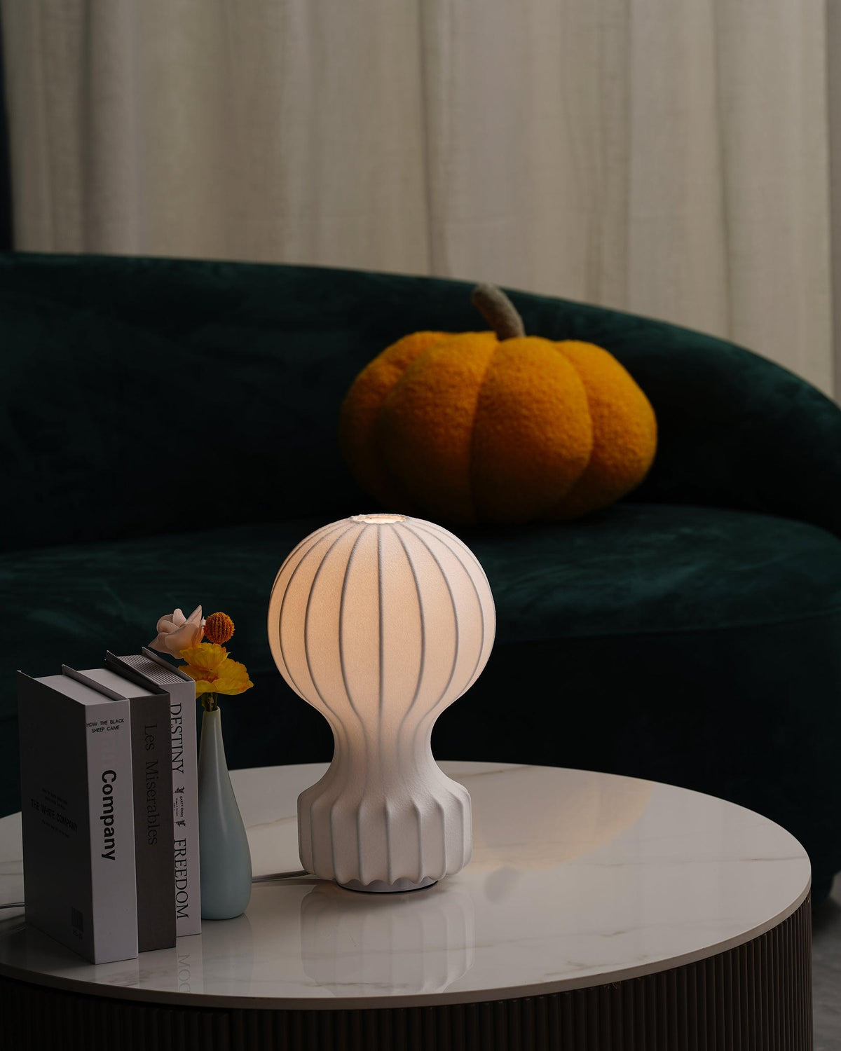 A lit Clérachic lamp on a round marble table in front of a green sofa, with books and a small vase at its side.