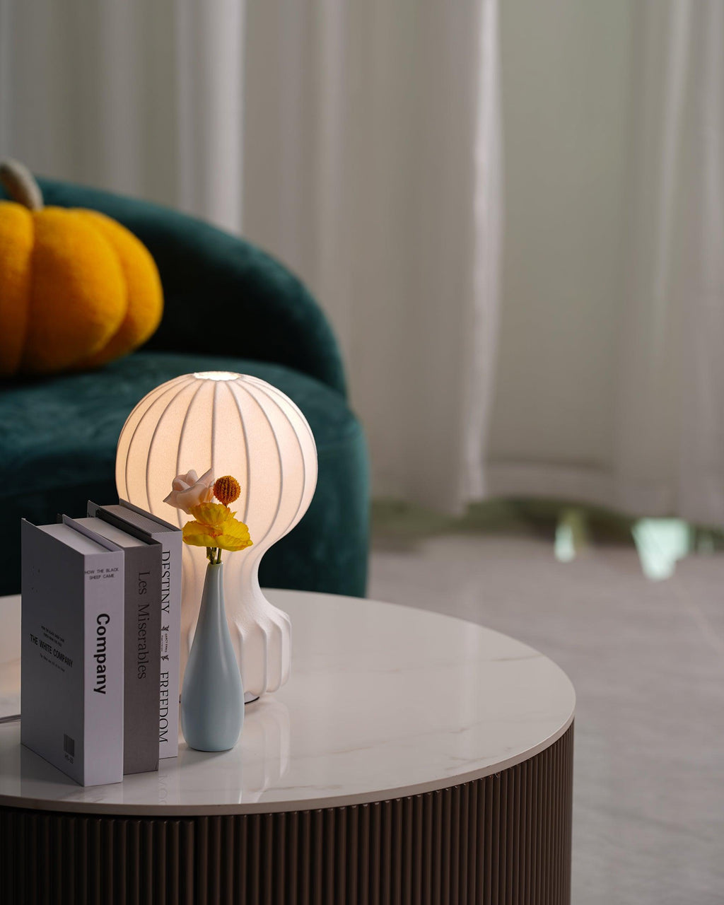 Clérachic lamp softly lit behind a pile of books and a blue vase, on a round marble table with a ribbed wooden base.