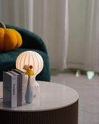 Clérachic lamp softly lit behind a pile of books and a blue vase, on a round marble table with a ribbed wooden base.