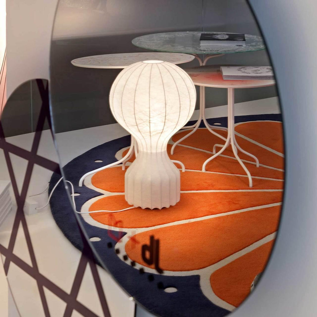 Lit Clérachic table lamp reflected in a round mirror, standing on an orange patterned rug beside a small marble-topped side table.