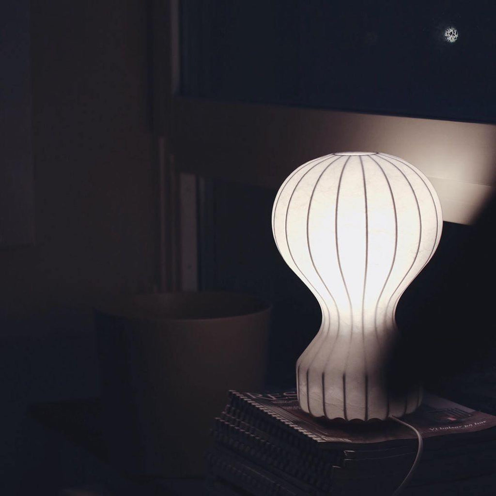 A Clérachic table lamp glowing softly in a dark room, its ribbed white shade resting on a stack of books.