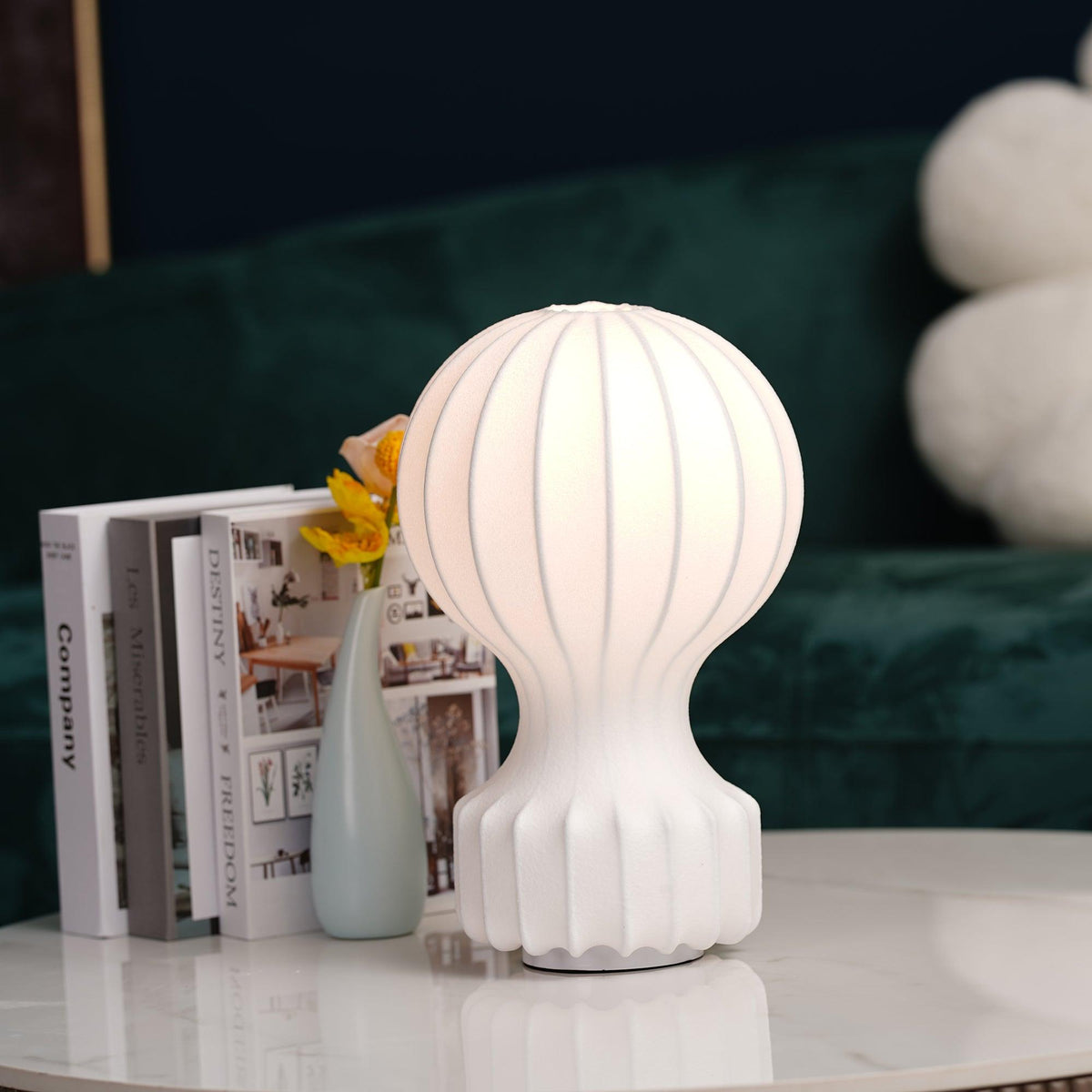 Lit white Clérachic lamp on a marble table in front of a green velvet sofa, with books and a slim pale-blue vase beside it.