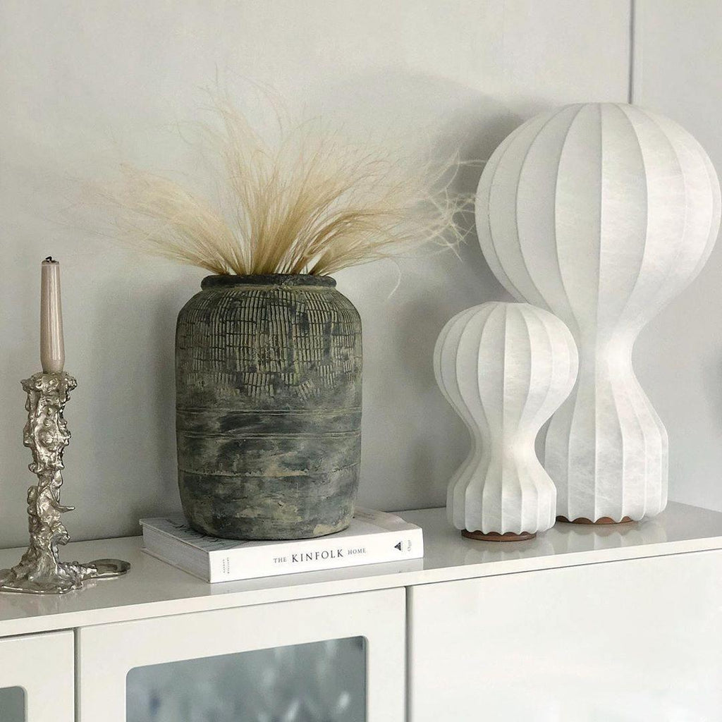 Two sizes of this Clérachic lamp side by side on a white cabinet, with dried grasses in a weathered stone vase and a candlestick nearby.