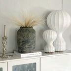 Two sizes of this Clérachic lamp side by side on a white cabinet, with dried grasses in a weathered stone vase and a candlestick nearby.