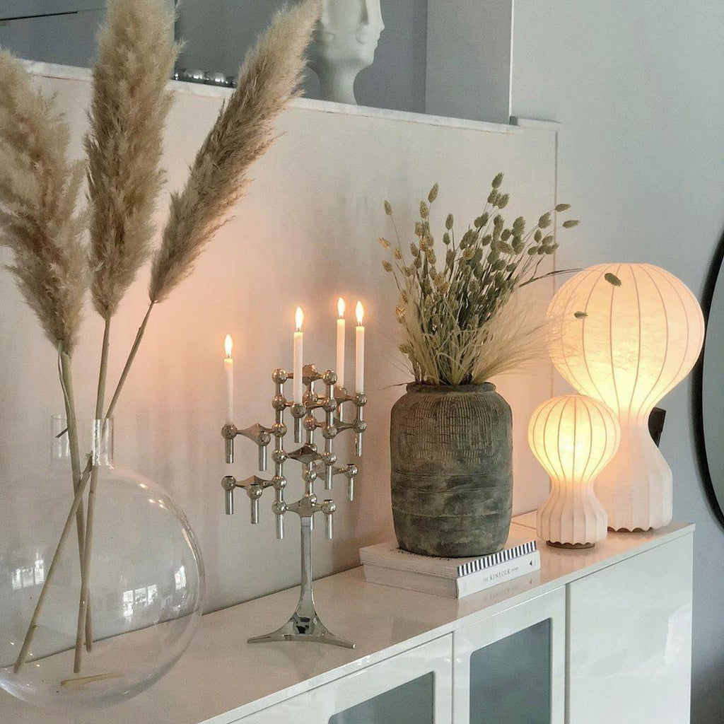 Both sizes of the Clérachic lamp glowing on a white sideboard, beside pampas grass, dried flowers and a silver candle holder.