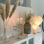 Both sizes of the Clérachic lamp glowing on a white sideboard, beside pampas grass, dried flowers and a silver candle holder.