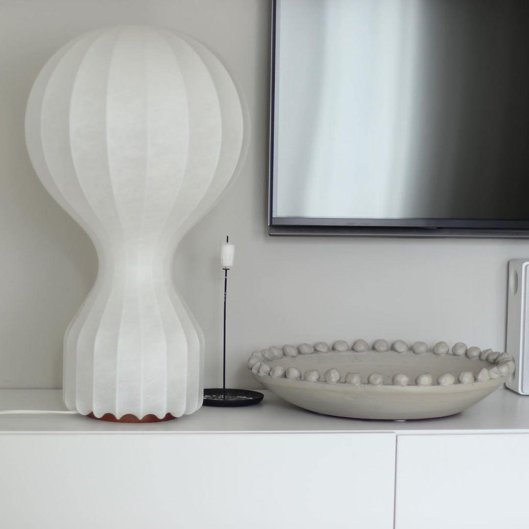Tall Clérachic lamp on a white sideboard beneath a wall-mounted TV, next to a beaded stone bowl and a slim black candlestick.