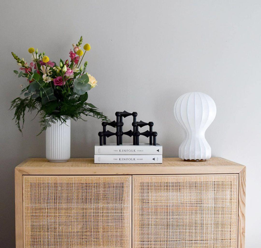 Small Clérachic table lamp on a rattan-fronted oak cabinet, beside a vase of spring flowers and a black candle holder.