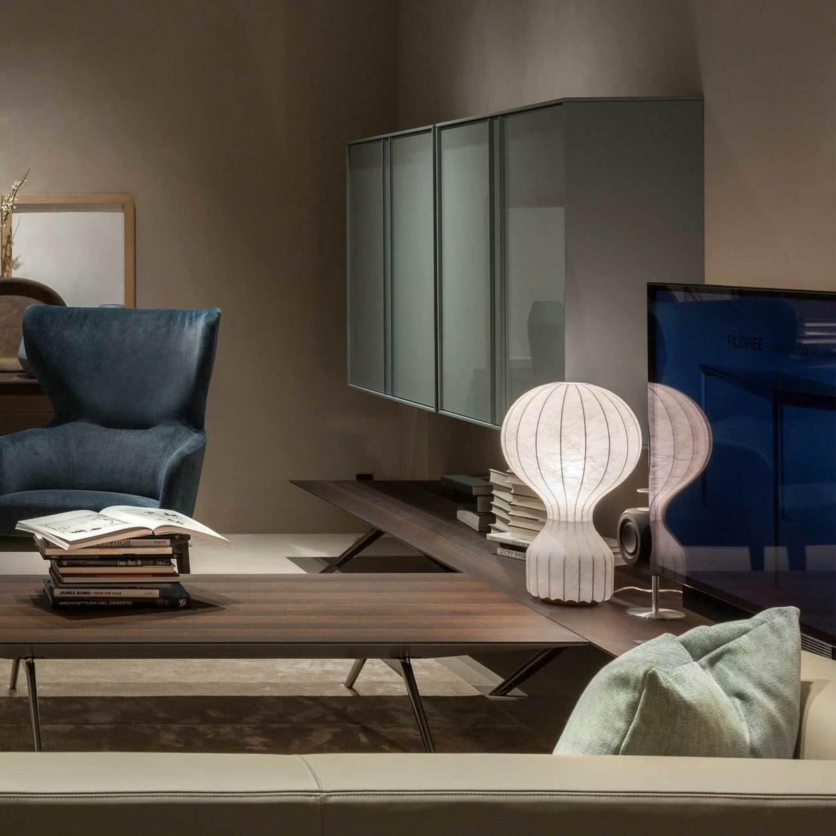 A Clérachic lamp lit on a low wooden table in a warm taupe living room, with a blue armchair and books close by.