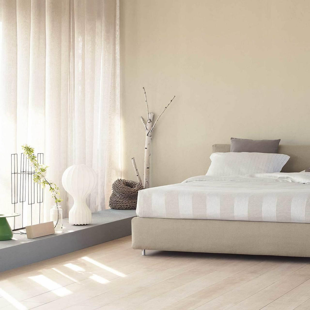 Clérachic table lamp on a low plinth beside a bed in a sunlit bedroom, with sheer curtains and pale wooden floorboards.