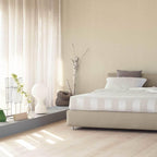 Clérachic table lamp on a low plinth beside a bed in a sunlit bedroom, with sheer curtains and pale wooden floorboards.