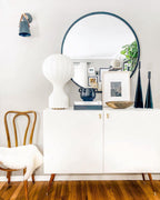 White Clérachic lamp on a white sideboard with tapered legs, in front of a round black-framed mirror and a framed print.