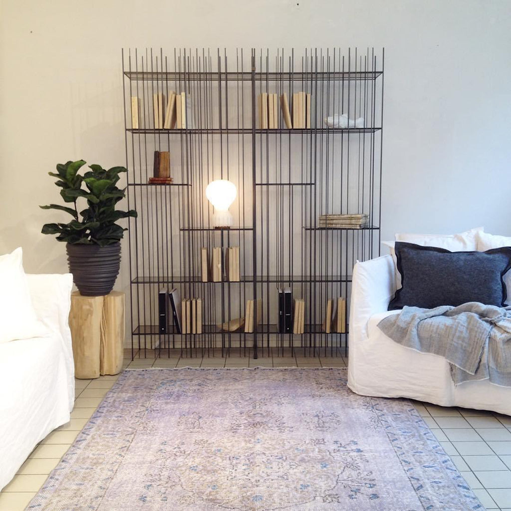 A small Clérachic lamp glowing on a shelf of a black metal bookcase, in a white room with a fiddle-leaf fig and a patterned rug.