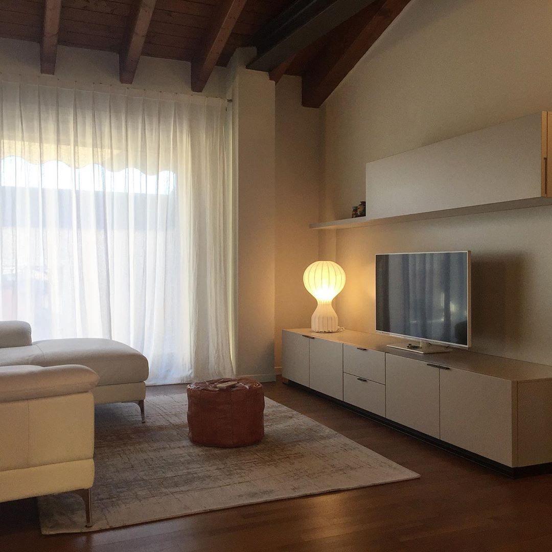 Distant view of a lit Clérachic table lamp on a pale TV unit, in a living room with sheer curtains and a leather pouffe.