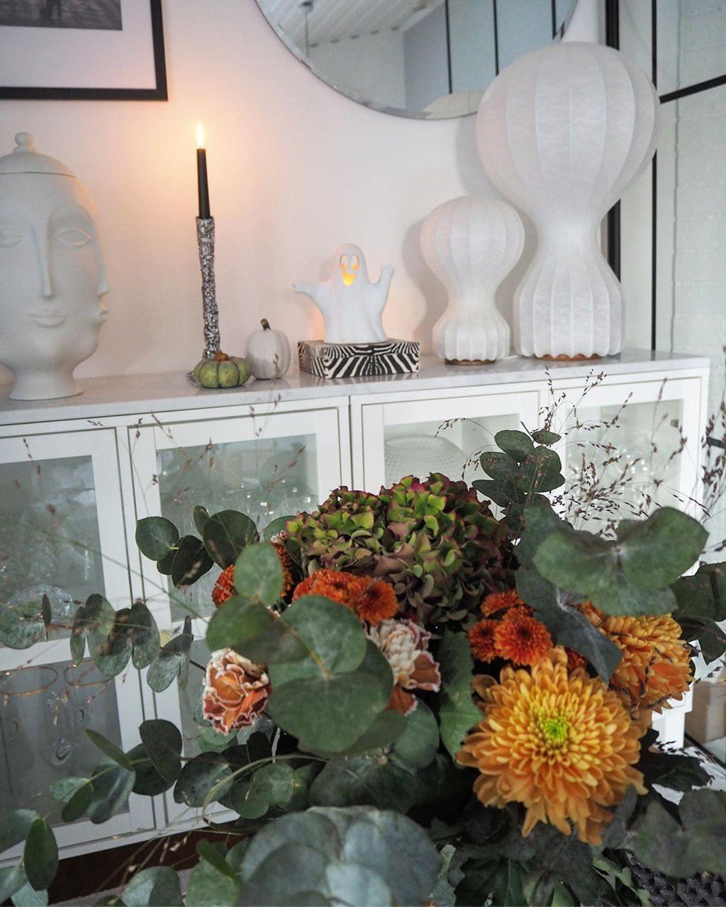 Two sizes of the Clérachic lamp on a white glazed cabinet, with a lit candle, a sculpted white head and an autumn flower arrangement in front.