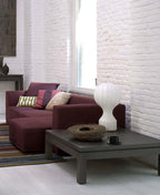 A Clérachic table lamp on a low dark table, with a bird's-nest bowl, in front of a maroon sofa and a white brick wall.