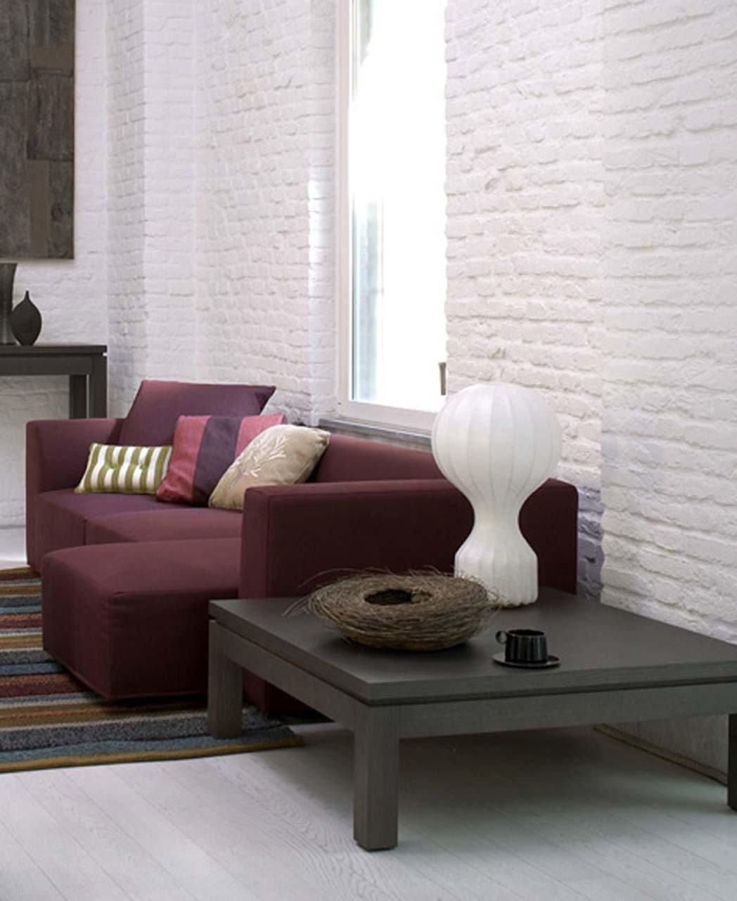 A Clérachic table lamp on a low dark table, with a bird's-nest bowl, in front of a maroon sofa and a white brick wall.
