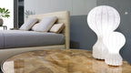 Two Clérachic lamps lit on a glass-topped round table in front of a bed with grey cushions.